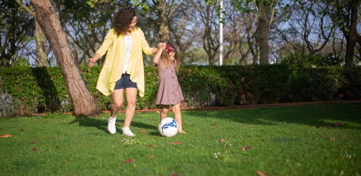A mother and daughter enjoying a sunny day playing soccer in a lush green park.