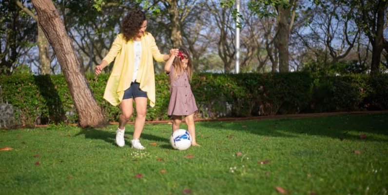 A mother and daughter enjoying a sunny day playing soccer in a lush green park.