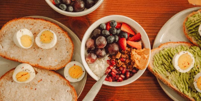 A fresh and vibrant breakfast flat lay featuring a yogurt bowl with fruits and toast with eggs and avocado.
